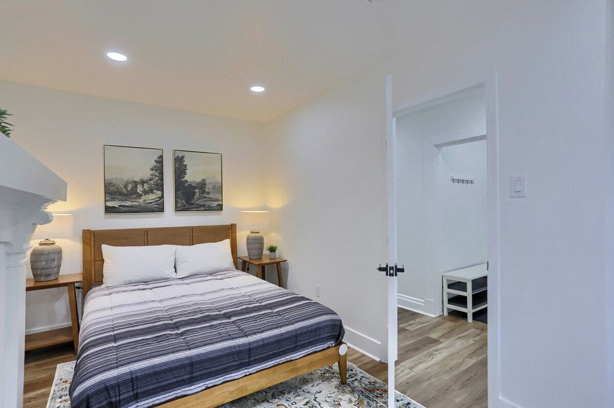 Een slaapkamer met een bed, twee aan de muur hangende foto’s, een nachtkastje met een lamp, een kleine plant en een deur die naar een andere kamer leidt.