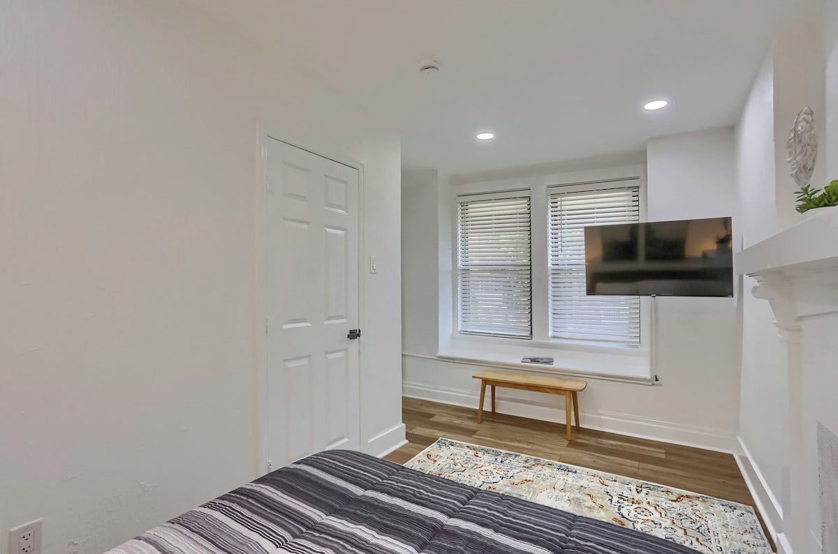 A bedroom with a bed, a bench, a TV, and a window with blinds.