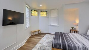 Un dormitorio con cama, televisión, banco, dos ventanas con persianas y mesita de noche con lámpara.