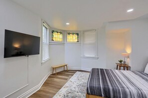 Un dormitorio con cama, televisión, banco, dos ventanas con persianas y mesita de noche con lámpara.