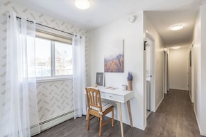 A room with a desk, chair, window with curtains, a framed picture, and a hallway.