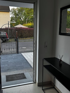 An open door leading to a paved area with a metal gate, a parked car, and a hedge in the background. - Guesthouse Cologne North For business travelers, tourists & families (Köln)