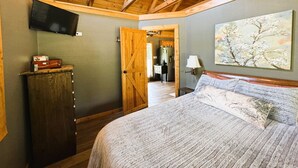 Une chambre avec un plafond en bois, un lit avec une literie à motifs, un téléviseur fixé au mur et un tableau.