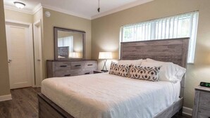 Une chambre à coucher avec un grand lit, une commode, un miroir, une table de chevet et une fenêtre avec des stores.