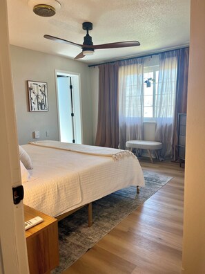 Quarto com cama, ventoinha no teto, janela com cortinas, mesa de cabeceira e uma cadeira.