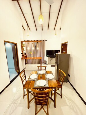 Een eethoek met een houten tafel voor vier personen, stoelen, en een keuken op de achtergrond, plus een hangende lamp.