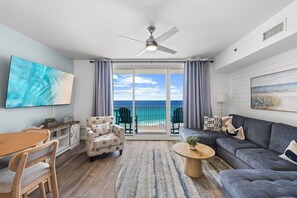 A living room with a sofa, armchair, and dining table, a ceiling fan, and a balcony with a view of the ocean. - 3bd Beachfront Resort Condo| Pools, Hot Tubs & Views (Panama City Beach)
