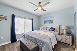 A bedroom with a bed, a nightstand, a lamp, a ceiling fan, a window with curtains, and a painting on the wall. - 3bd Beachfront Resort Condo| Pools, Hot Tubs & Views (Panama City Beach)