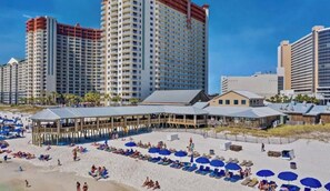 Un hotel frente a la playa con varios edificios altos, una zona de playa cubierta con sillas y sombrillas de descanso, y una playa de arena.