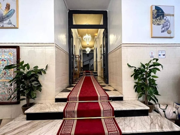 Eine Hotelhalle mit einer Marmortreppe, einem roten Teppich, Topfpflanzen und Kunstwerken an den Wänden.