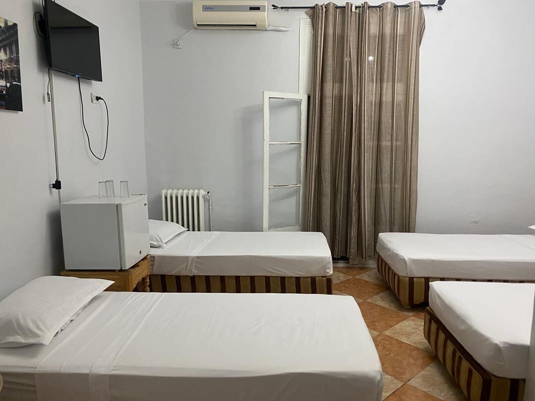 Habitación de hotel con dos camas individuales, un armario blanco pequeño, un radiador, un televisor montado en la pared y una ventana con cortinas.