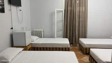 Habitación de hotel con dos camas individuales, un armario blanco pequeño, un radiador, un televisor montado en la pared y una ventana con cortinas.
