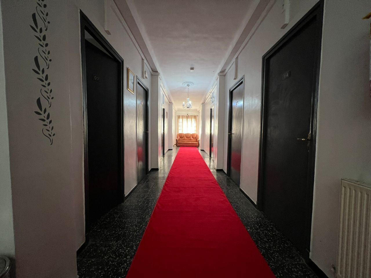 A hotel hallway with a red carpet, doors on both sides, and a chandelier in the distance.