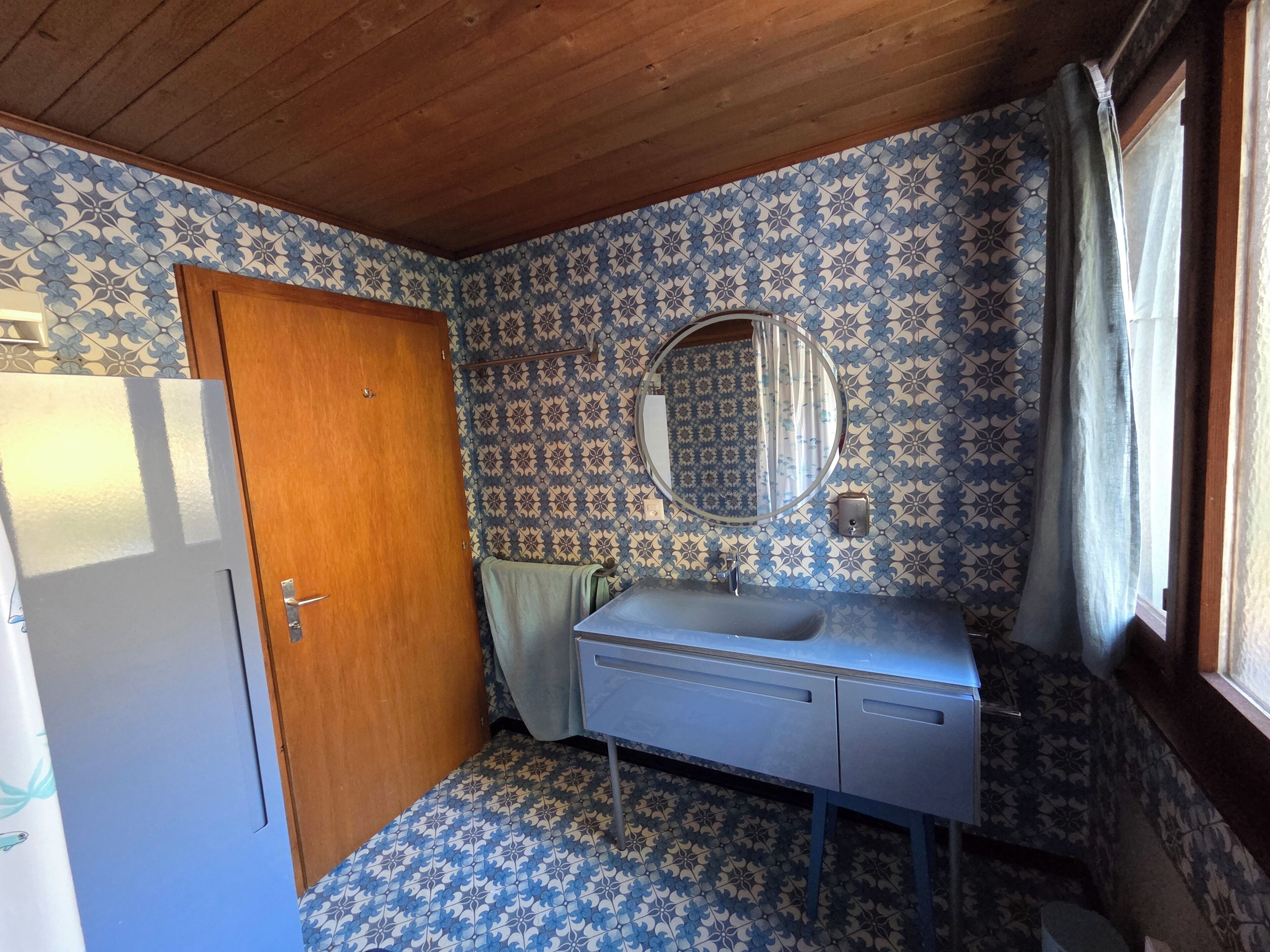 Un baño con paredes estampadas, una puerta de madera, un lavamanos con espejo y una ventana con cortinas.