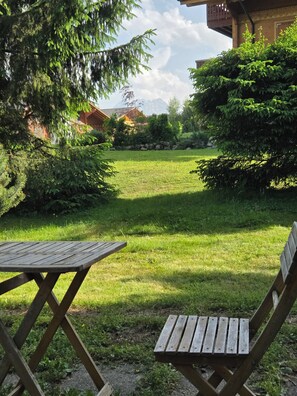 Un espacio al aire libre con césped, una mesa y sillas de madera, rodeado de árboles y un edificio al fondo.