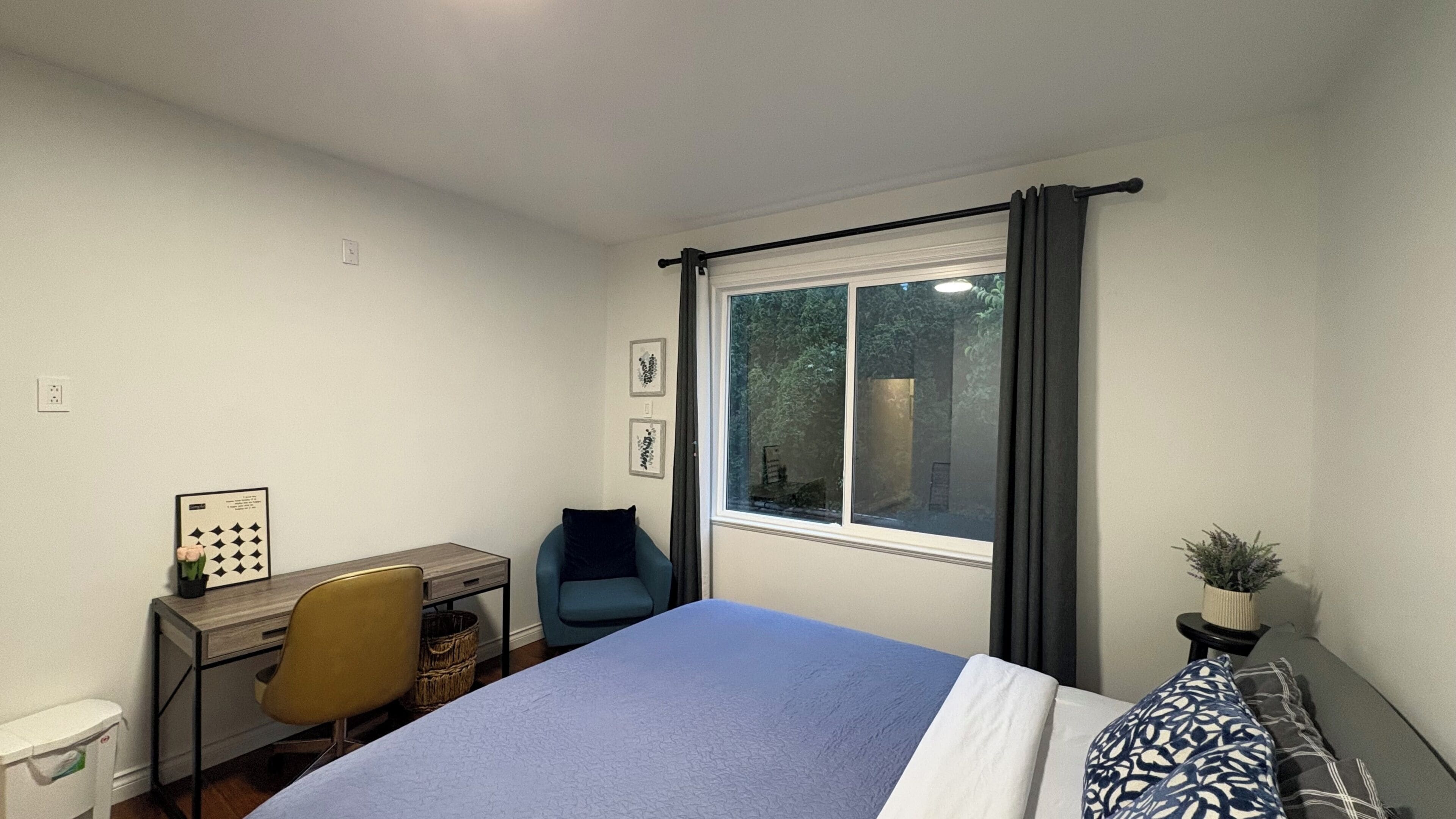 A bedroom with a bed, a desk, a chair, a window with curtains, and a small plant.