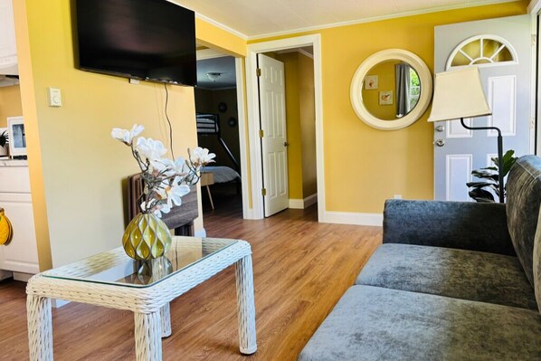 A living room with a sofa, a glass coffee table, a TV mounted on the wall, and a door leading to another room.