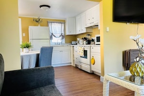 A compact kitchen with white cabinets, a refrigerator, a microwave, and a stove. There is a dining table with chairs and a small table with a decorative vase.