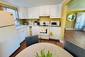 A compact kitchen with white cabinets, a microwave, a stove, and a refrigerator. There is a dining table with chairs and a potted plant.