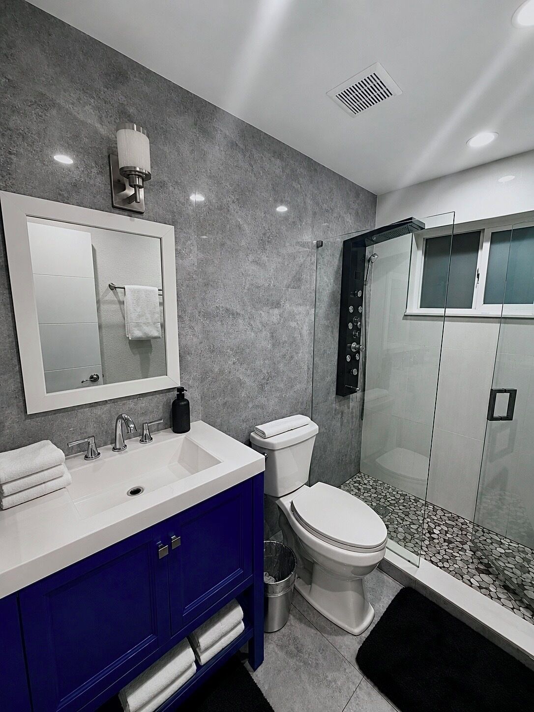 A modern bathroom with a large mirror, a white sink, a blue vanity, a toilet, and a glass-enclosed shower.