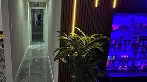 A hallway with a potted plant, a television displaying a cityscape, and a wooden panel wall with vertical lights. - Modern Retreat Near Beach, Las Olas & Hard Rock (Fort Lauderdale)