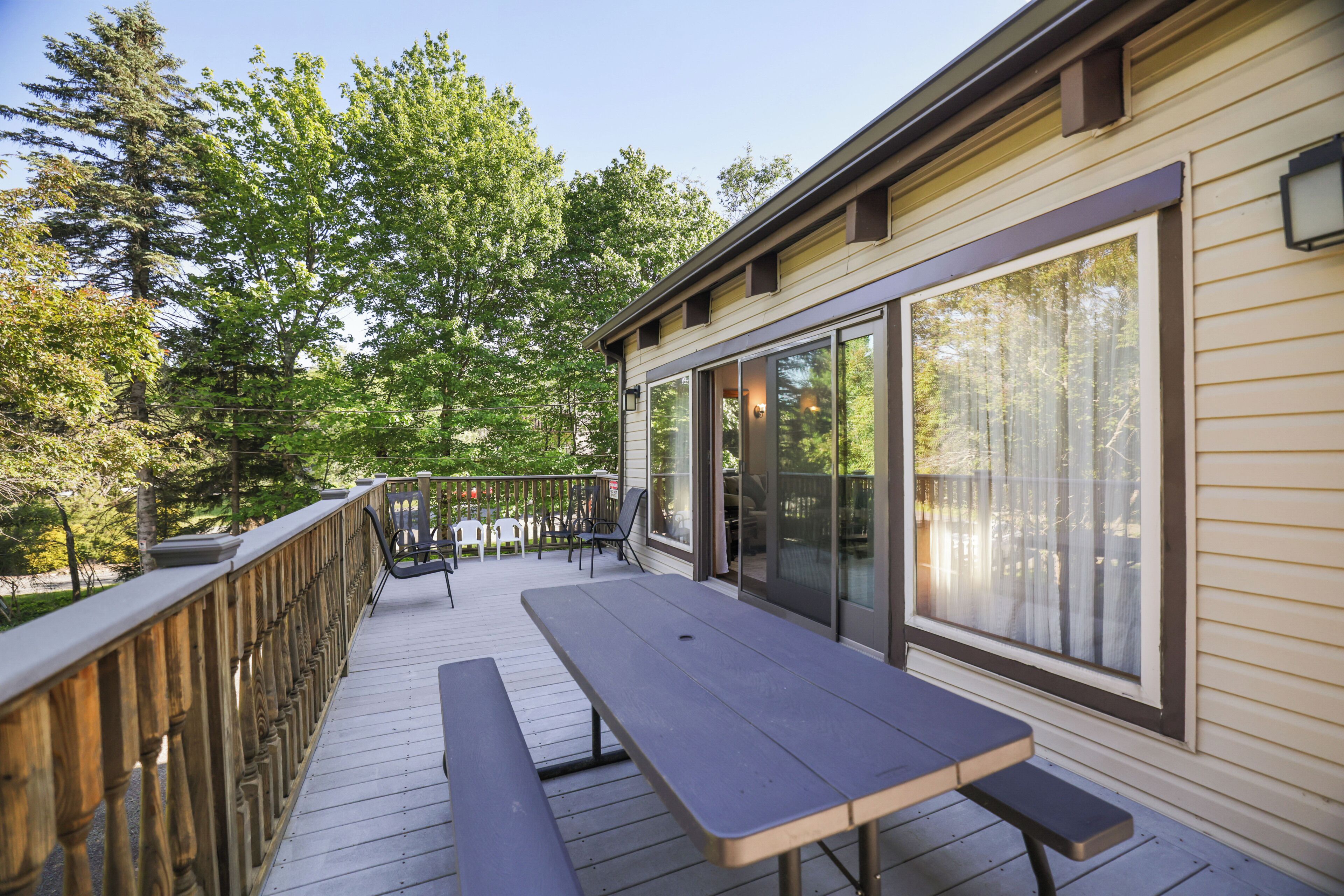 A wooden deck with a picnic table and chairs, surrounded by glass doors and windows, with a view of trees.