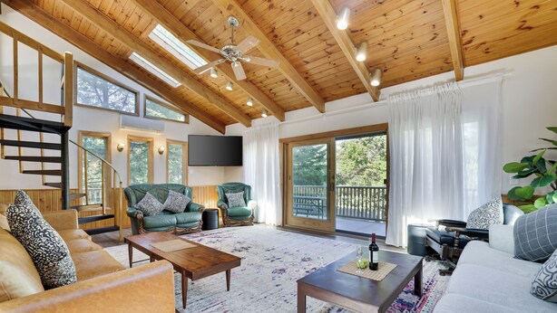 A spacious living room with wooden beams, a ceiling fan, a large TV, and a balcony with a view of trees.