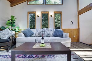 A living room with a sofa, a coffee table with a bottle and glasses, and a large plant. - Poconos Lake Cabin Retreat - Hot Tub, Sauna, Movie & Game Room (Albrightsville)