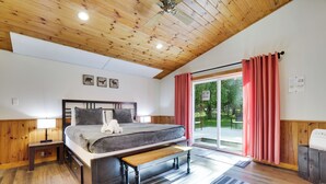 A bedroom with a wooden ceiling, a bed with a bench, a nightstand, a window with curtains, and a view of the outdoors.