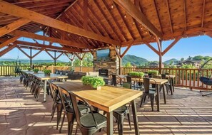 An outdoor patio with wooden tables and chairs, a stone fireplace, and a view of the mountains. - Amazing BRAND NEW 8bdrm Cabin-Pool-Arcades-Gazebo (Sevierville)