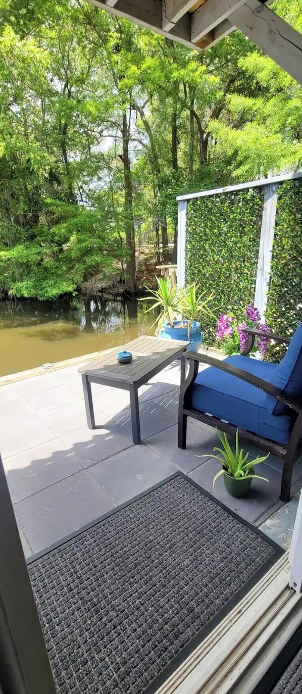 Un espacio de estar al aire libre con techo, con una mesa y sillas, rodeado de vegetación y un cuerpo de agua.