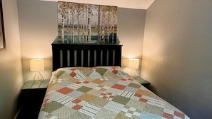 A bed with a quilt, two bedside lamps, a wall-mounted artwork, and a dark headboard.