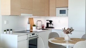 Une cuisine moderne avec des meubles en bois, un four encastré, une machine à café et un coin repas comprenant une table blanche et des chaises grises.