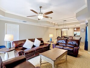 A living room with a brown leather sofa, a wooden coffee table, a ceiling fan, and a kitchen in the background. - Reunion Elegant Disney Condo, Resort Amenities (Kissimmee)