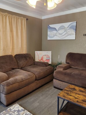 Ein Wohnzimmer mit zwei braunen Sofas, einem Holztisch, einem beigen Vorhang und einer Wand mit einem gerahmten Bild und einer Wanddekoration.