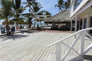 A wooden deck with palm trees, a thatched-roof structure, and a building with a sign. - Family 2BR Townhouse Beach Access, Pool & Fast Wi-fi (Punta Cana)
