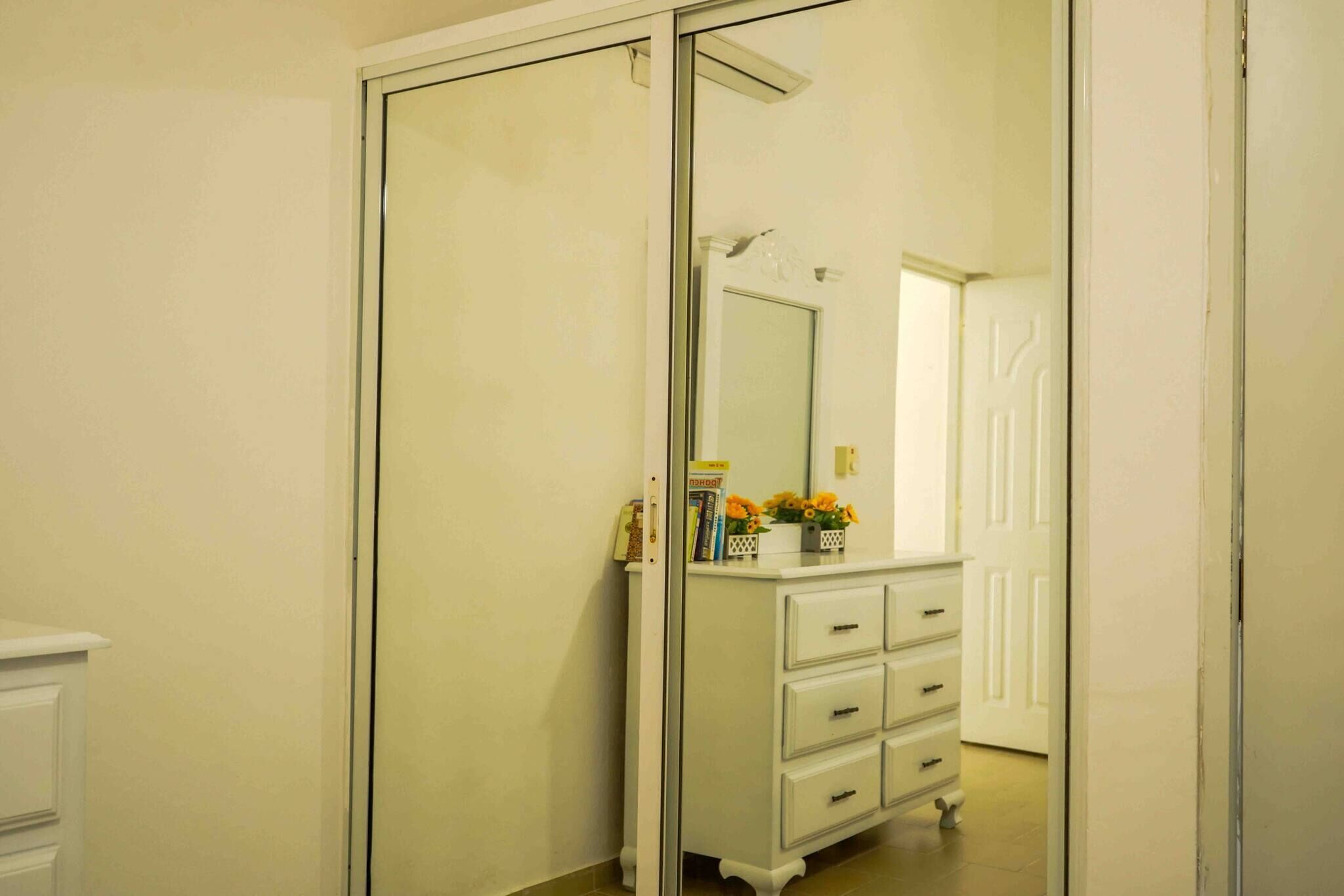 Een kamer met een schuifdeur, een witte dressoir met lades, een spiegel en een klein tafeltje met bloemen.