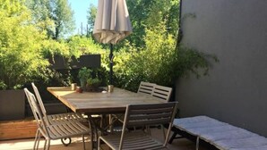 Terrassa exterior amb taula de fusta, caires metàl·liques, un parasol blanc i una baren verge coberta.
