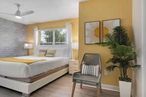 Une chambre à coucher comprenant un lit, des tables de chevet, une chaise, une fenêtre avec des rideaux, un ventilateur de plafond et des œuvres d’art murales.