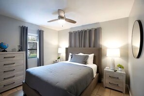 Ein Schlafzimmer mit Bett, Nachttisch, Kommode, Deckenventilator, einem Fenster mit Vorhängen und einem wandmontierten Spiegel.