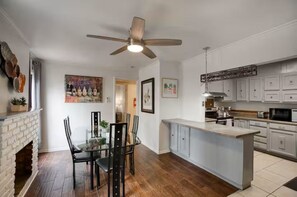 Eine Küche mit Kochinsel, weißen Schränken, Deckenventilator und Essbereich mit Kamin aus Ziegelsteinen.