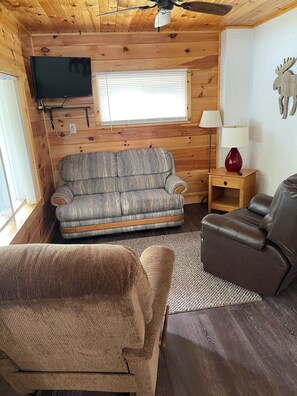 A cozy cabin interior with a sofa, a recliner, a TV mounted on the wall, and a ceiling fan.