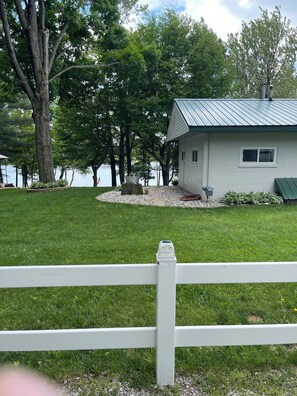 A house with a metal roof, surrounded by a white fence, near a body of water with trees.