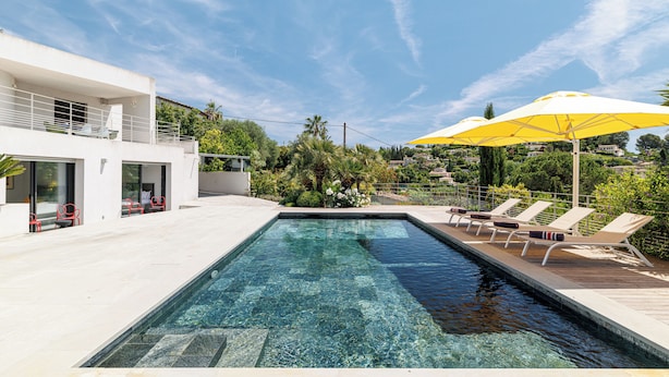 Ein rechteckiger Pool mit klarem Wasser, umgeben von einem weißen Gebäude mit Balkonen, roten Stühlen und einem gelben Sonnenschirm.
