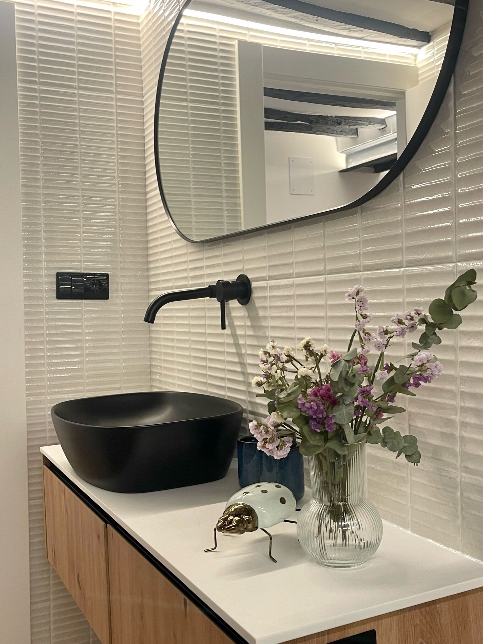 Un baño con un lavabo negro tipo cuenco, un espejo, un florero con flores y un insecto decorativo.