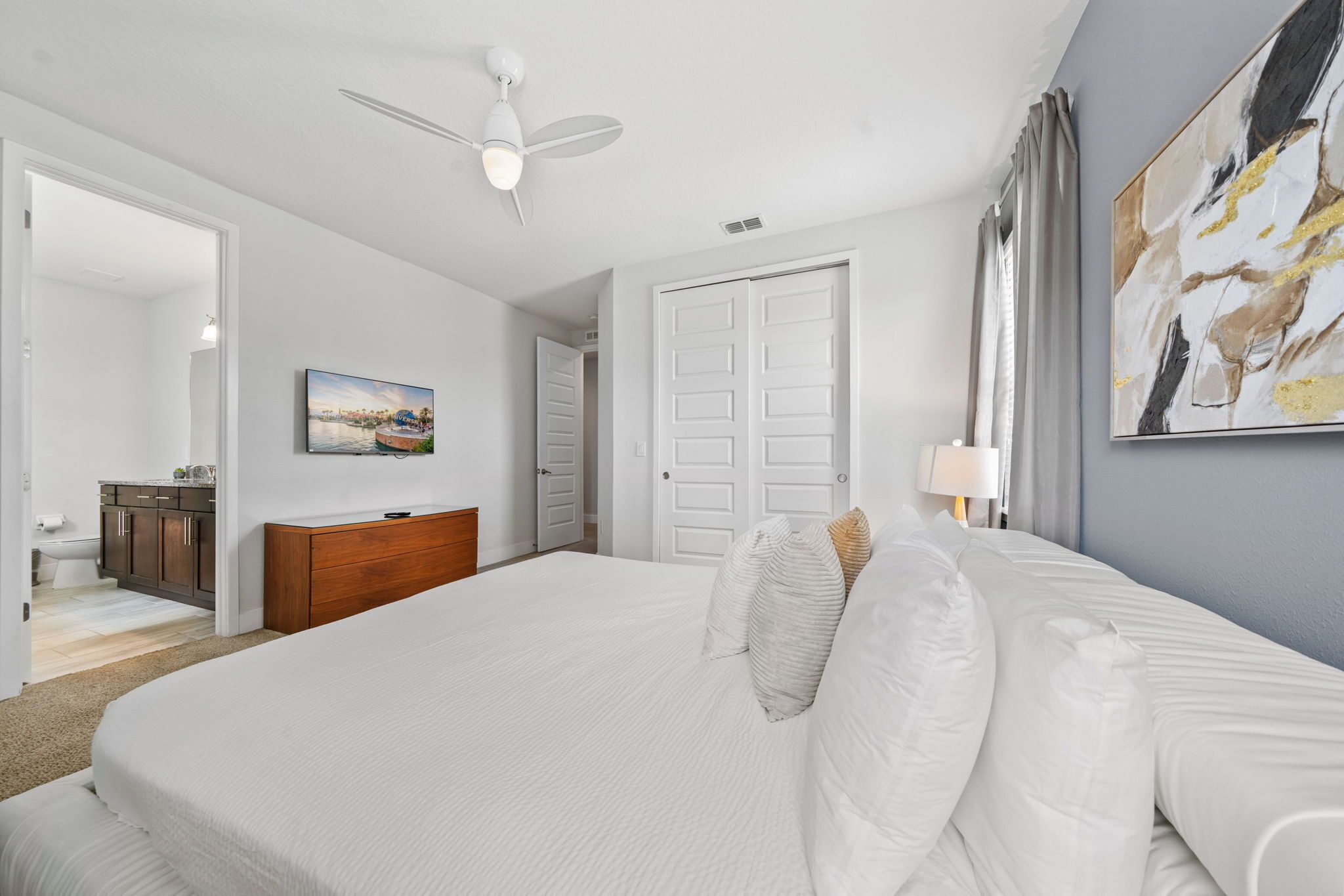 A bedroom with a large bed, a ceiling fan, a painting, a door, and a nightstand with a lamp.