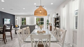 A modern dining area with a glass table, white chairs, a woven pendant light, and a view of the living room. - 9Br Spacious Villa With Private Pool Close Disney (Kissimmee)