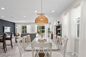 A modern dining area with a glass table, white chairs, a woven pendant light, and a view of the living room. - 9Br Spacious Villa With Private Pool Close Disney (Kissimmee)