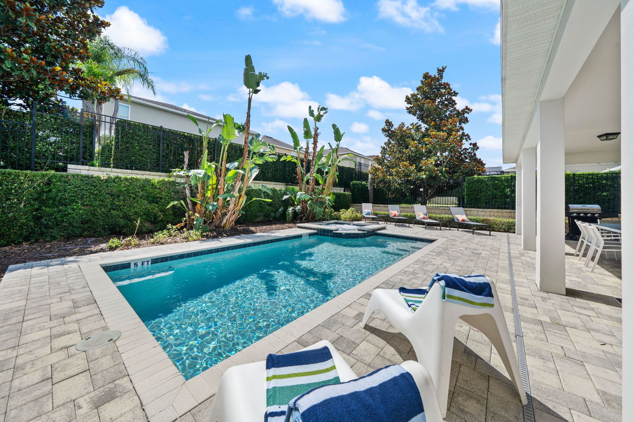 Une piscine entourée de chaises longues, d’une terrasse dallée et de verdure.