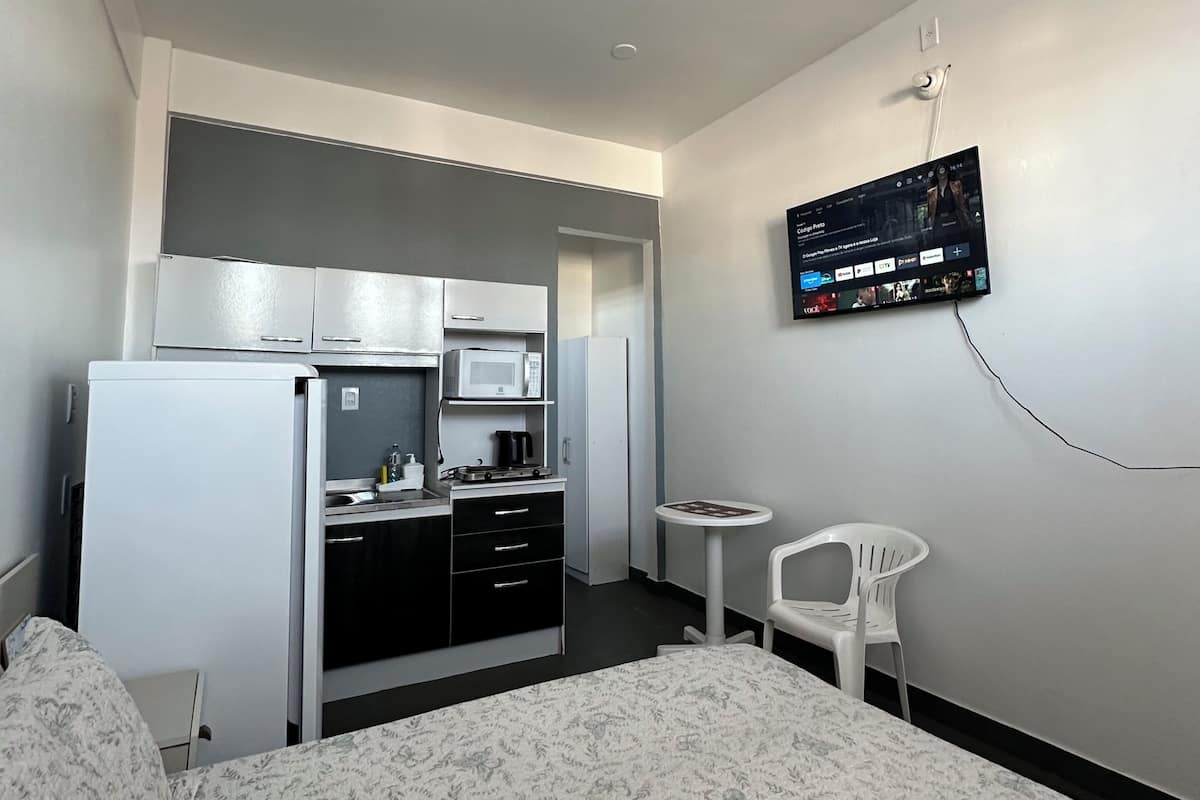 A hotel room with a bed, a TV mounted on the wall, a kitchenette, and a small table with a chair.